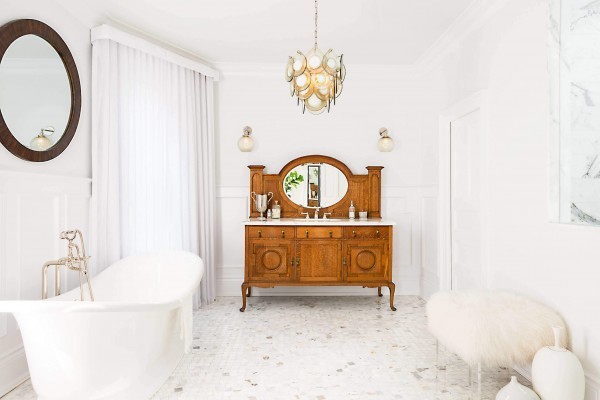 White bathroom with free standing tub, white marble mosaic floor, wood vanity with mirror, glass globe sconces, fur bench, white drapes, pendant light, and round wood mirror White bathroom with free standing tub, white marble mosaic floor, wood vanity with mirror, glass globe sconces, fur bench, white drapes, pendant light, and round wood mirror