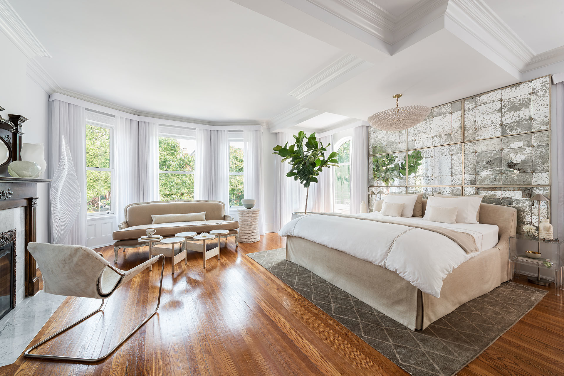 bedroom with distressed mirrored wall, beige bed, acrylic side tables, chrome floor lamp, ficus, settee, white floor to ceiling sheers, hardwood floor, oversized pendant light, and white marble fireplace bedroom with distressed mirrored wall, beige bed, acrylic side tables, chrome floor lamp, ficus, settee, white floor to ceiling sheers, hardwood floor, oversized pendant light, and white marble fireplace